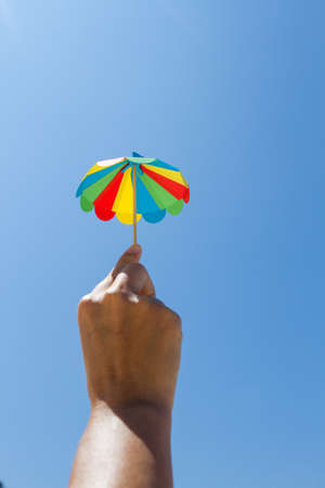 small colored paper cocktail umbrella on the beach, holding in his hand on blue sky backgroundの写真素材