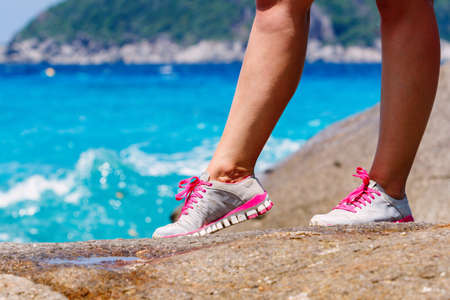 Runner woman stands on a rock near the blue sea. Red laces gray sneakers. Closeup. Photo kneeの写真素材