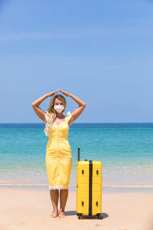 A woman in a protective surgical mask on her face, walking along the beach with a yellow suitcase. coronavirus disease COVID-19 is a dangerous virus. Pandemicの写真素材