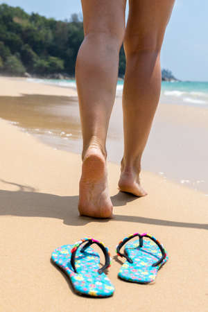 Girl rear view walking barefoot on the sand. Beach shoes in the foreground.