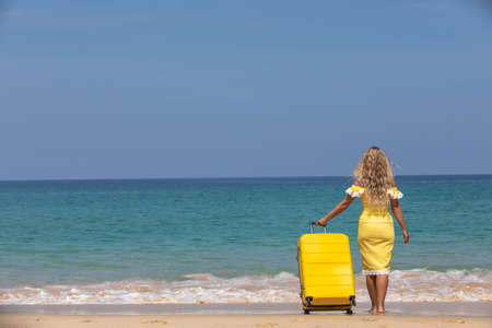 Beautiful girl in a yellow dress on the beach with a yellow suitcase. Blue sea. Yellow sandの写真素材