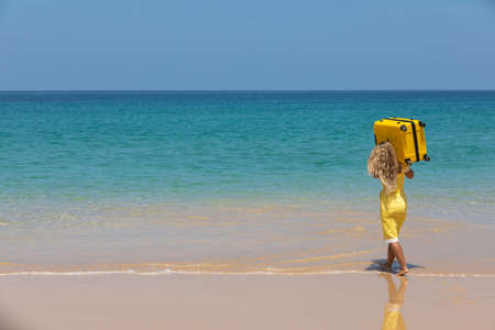 Beautiful girl in a yellow dress on the beach with a yellow suitcase. Blue sea. Yellow sandの写真素材