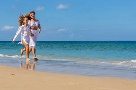 Young beautiful mother with her charming daughter in white clothes walk on a sandy beach near the waterの写真素材