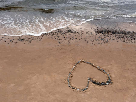 The heart is lined with small pebbles on the sand on the seashore and the oncoming sea wave