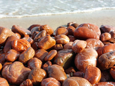 Unique red jasper beach. Many pieces of red jasper polished by the sea. Wet sea stones on a blurred background of the oncoming wave.の写真素材