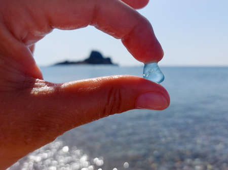 A piece of blue glass rolled by the sea in two fingers against a blurred background of the sea and a small island.の写真素材