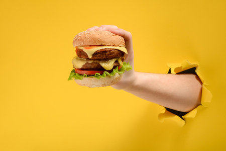 Hand holding a big burger through a hole in torn yellow paper wallØ Special offer and cheap price on fast foodの写真素材