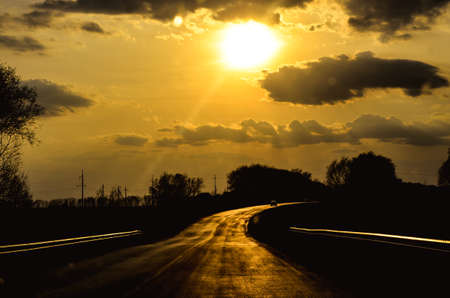 The road in the rays of a bright sunset. In the sky above the road, the silhouette of dark cloudsの写真素材