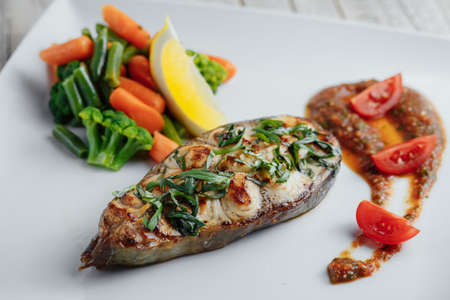 Baked fish on a white plate with tomatoes, carrots and herbs on a wooden tableの写真素材