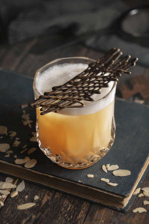 Whiskey sour cocktail with foam on an old book on a wooden surface with a chocolate net and almond shavingsの写真素材