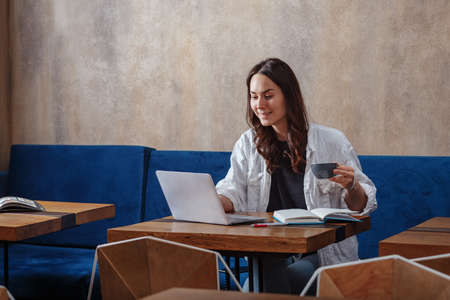 A young girl smiling nicely sitting in a cafe and working on her laptop, drinking coffee, keeping notes in a notebookの写真素材