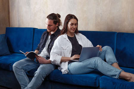A young couple spends time at home, studying and playing or working, reading, receiving new information from a laptop while sitting on a sofa. Cheerful couple chooses a movie on a laptop, relaxing together at the weekend at home. Remote work. Work from homeの写真素材