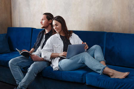 Quarantined life. A young couple spends time at home, studying and playing or working, sitting on the couch. Cheerful couple chooses a movie on a laptop, relaxing together at the weekend at home. Remote work. Work from homeの写真素材