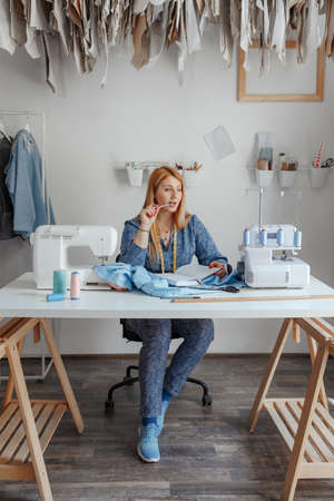 Portrait of happy young lady fashion designer smiling standing in workshop alone looking at camera. Small business owner and successful youth concept. You can see the thread, sketches, the fabric and the tool of the tailorの写真素材