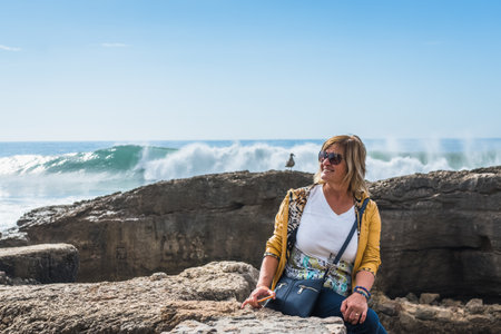 A middle-aged woman sits on the shore of the Atlantic Ocean, watching the waves crash against the shore and create a white splash. Blue pants, yellow jacket and sunglasses. Blue skの写真素材