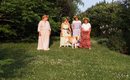 Four women, adorned with homemade wreaths, stand in the garden on a summer solstice evening, with a Labrador next to them.の写真素材