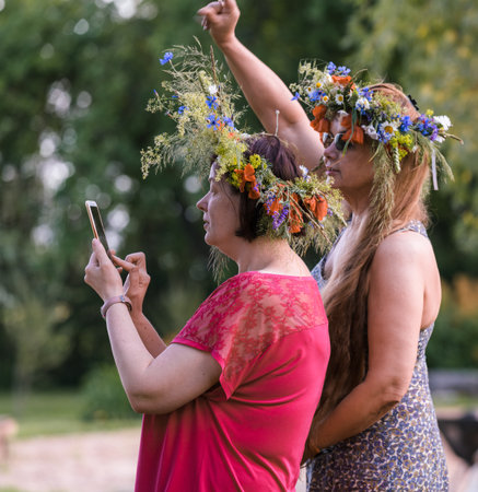 Two women with summer solstice wreaths on their heads stand side by side and look at the phone held by the one in the red dress.の写真素材