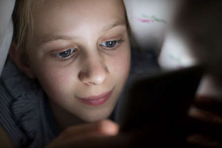 Girl Using Mobile Phone Hiding Under Duvet In Bed At Night