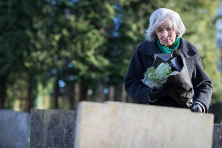 Sad Senior Woman With Flowers Standing By Graveの写真素材