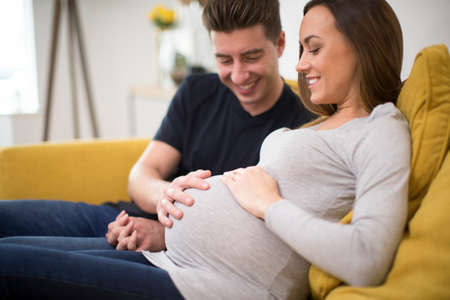 Pregnant Couple Sitting On Sofa With Man Touching Womans Stomach
