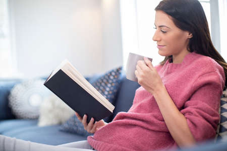 Woman With Hot Drink Relaxing On Sofa At Home Reading Bookの写真素材