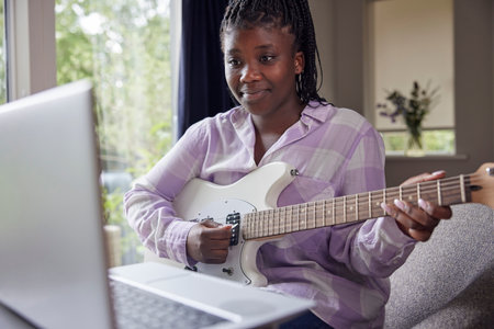 Teenage Girl At Home Learning To Play Electric Guitar With Online Lesson On Laptop Computerの写真素材