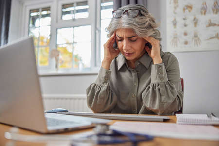 Stressed And Overworked Mature Female GP At Desk In Doctors Officeの写真素材