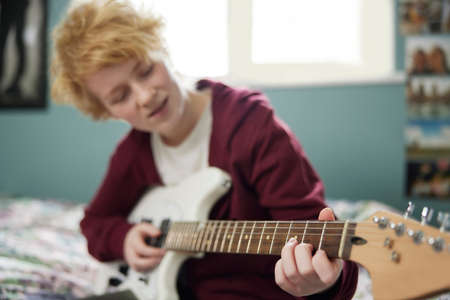 Teenage Girl Sitting On Bed Learning To Play Electric Guitar In Bedroomの写真素材