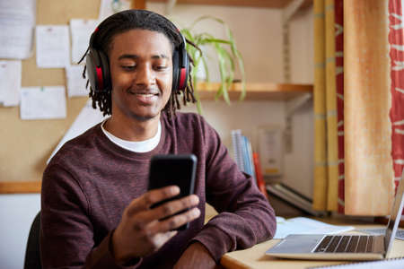 Male University Or College Student Working On Laptop Wearing Wireless Headphones Looking At Mobile Phone  At Desk In Roomの写真素材
