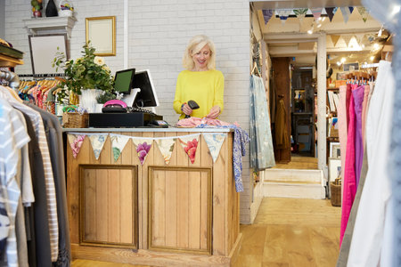 Female Volunteer Working In Charity Shop Or Thrift Store Selling Used And Sustainable Clothing And Household Goodsの写真素材
