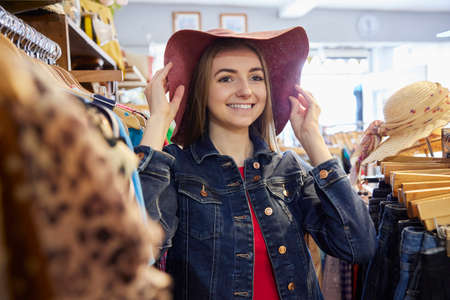 Young Woman Buying Used Sustainable Clothes From Second Hand Charity Shop Trying On Hatの写真素材
