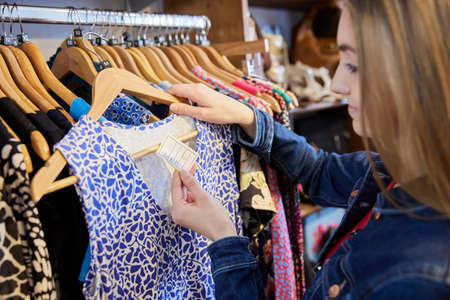 Young Woman Buying Used Sustainable Clothes From Second Hand Charity Shop Looking At Price Tagの写真素材