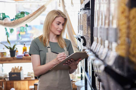 Female Owner Of Sustainable Plastic Free Grocery Store Checking Stock On Shelves
