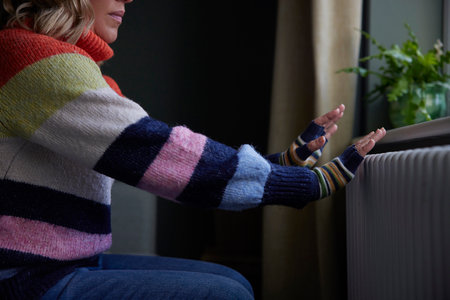 Woman At Home In Gloves Warming Hands By Radiator During Cost Of Living Energy Crisisの写真素材