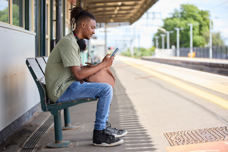 Young Man Commuting To Work On Train Sitting  On Platform Looking At Mobile Phone For Travel Information Or Social Mediaの写真素材