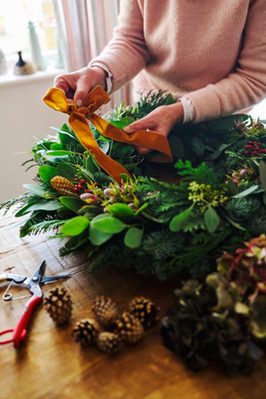 Close Up Of Woman Making And Decorating Festive Christmas Wreath On Table At Home Fastening Bow Made From Ribbonの写真素材