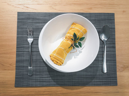 Top view yellow cotton napkin decorated with leaves with white plate, spoon and fork in grey placemat on wooden table.の写真素材