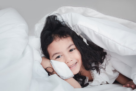 Little kid girl after sleeping lying in his white blanket in bed. Cute asian little girl happy playing and smiling on white background. Copy space for text.の写真素材