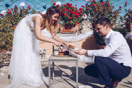 Beauty bride and handsome groom are cutting a wedding cake. Couple on the terrace with Rome cityscape in the background. Beautiful model girl in white dress. Man in suit.の写真素材