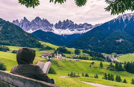 Tourist reading a book sitting on the wooden bench at autumn forest and mountains background, Santa Magdalena village, Dolomites, South Tyrol, Italyの写真素材