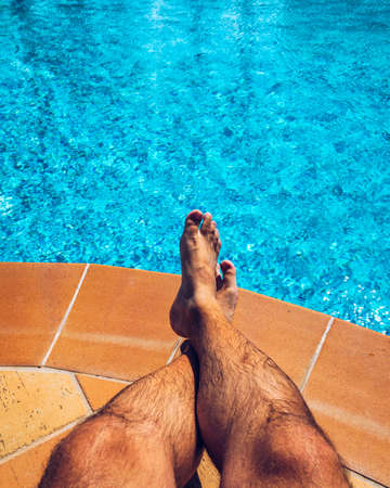 Man enjoying the hot summer at swimming-pool. Sunbathing by the swimming pool, man's legs lying down on a sun lounger over the water.の写真素材