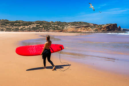 Unrecognizable young woman carrying surf board at Praia da Bordeira, Portugal.