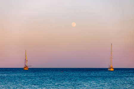 Full moon rising over the water with a small sailing boat in the foreground. Sailing boat with raising moon at sunset. Moon rising over the sea and yacht floating on the water surface. Sardinia, Italyの写真素材