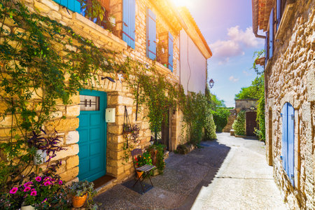 Charming narrow street in Venasque with sunlit stone and vibrant flowers, Provence, France. Narrow street in picturesque village of Venasque features stone buildings adorned with vibrant flowers.の写真素材