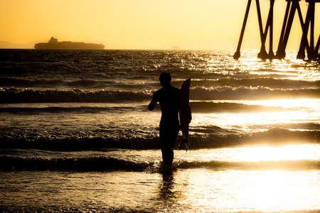 Silhouette of a surfer walking on the beach carrying a surfboard in front of the pier at Huntington Beach, California at sunsetの写真素材