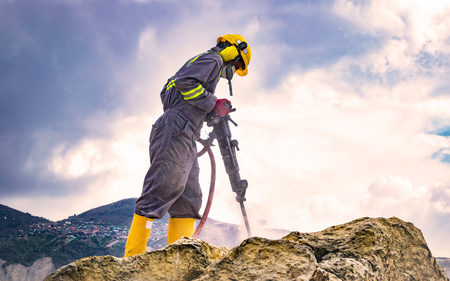 Worker with helmet and protective suit using a drilling machine on top of a large rockの写真素材
