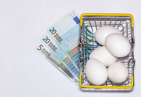 Fresh eggs in a shopping cart with various euro bills underneath it. Shopping, purchasing, and food delivery concept. White background. Close up shot. Isolated.の写真素材