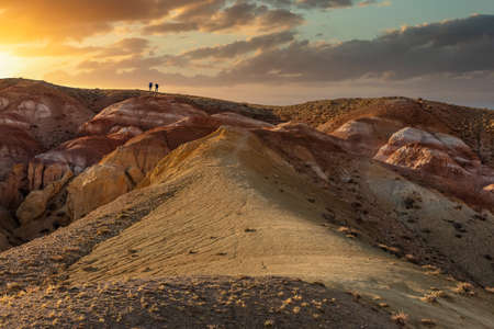 Beautiful low angle shot of two tiny human figures walking towards sunset on top of massive red...