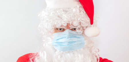 Close-up shot of man wearing a Santa Claus costume and a medical mask on white background. Man looks very angry and stressed, staring straight in the camera. New normal, new reality, holiday conceptsの写真素材