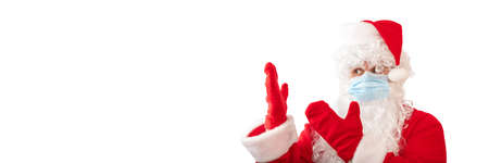 View of a man wearing a Santa Claus costume, medical mask and pointing with both hands at something next to him, isolated on white background. Banner size, copy space. Pandemic holiday concept.の写真素材